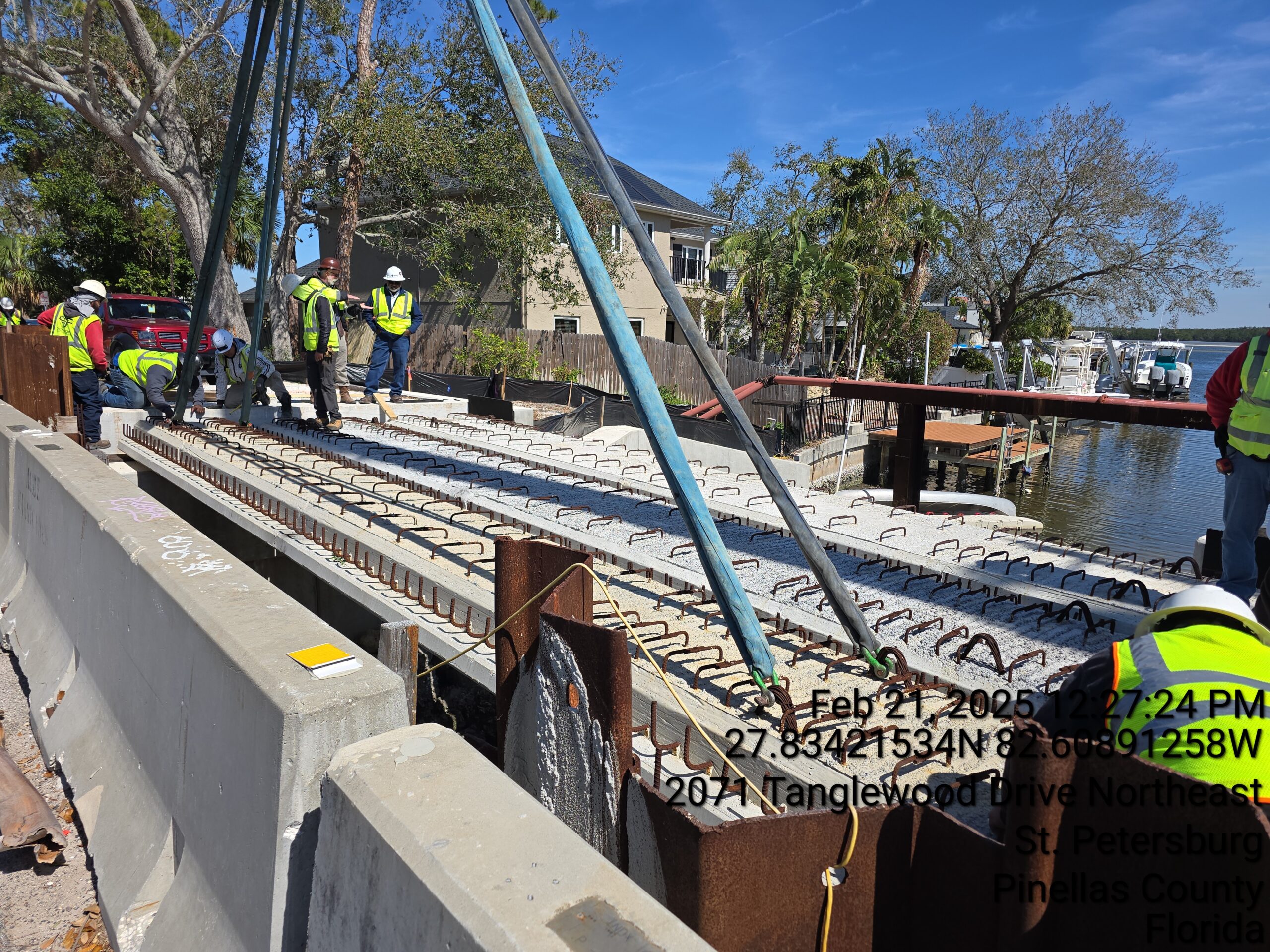 Construction workers install beams at a riverside site. Text: Feb 21 2025 12:27 PM 27.83421534N 82.60891258W 2074 Tanglewood Drive Northeast St Petersburg Pinellas County Florida