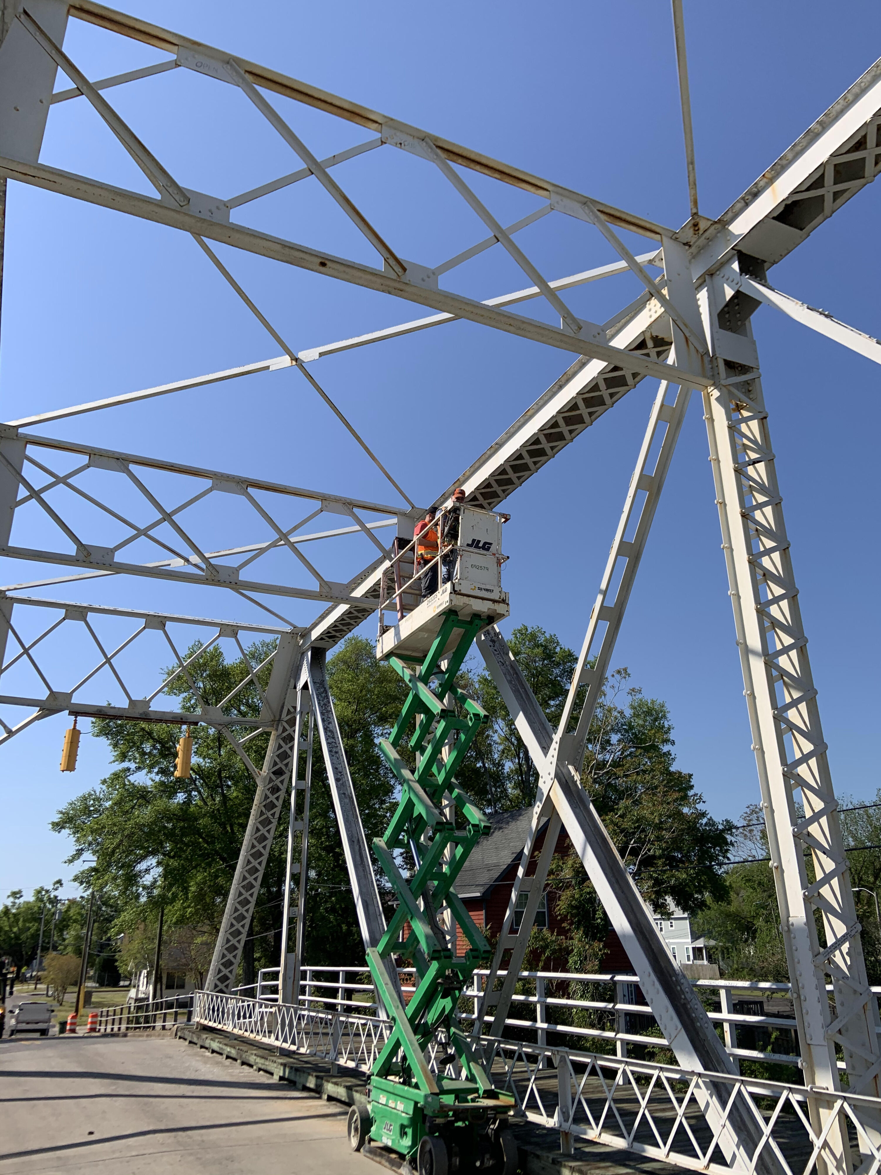 Worker on lift inspecting white metal bridge framework under clear blue sky
