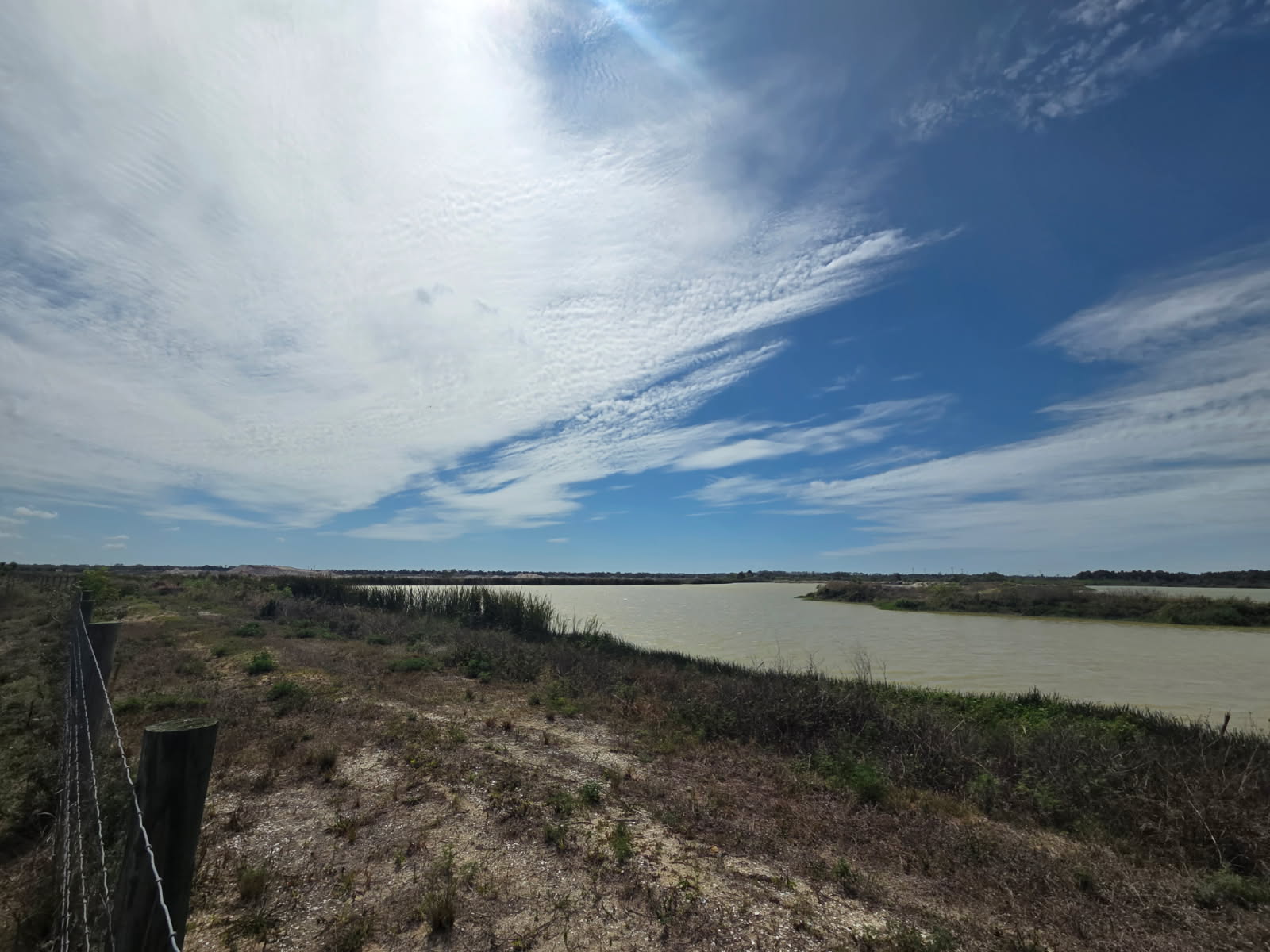 Vast cloudy sky over serene riverside landscape with greenery and a fence on the left