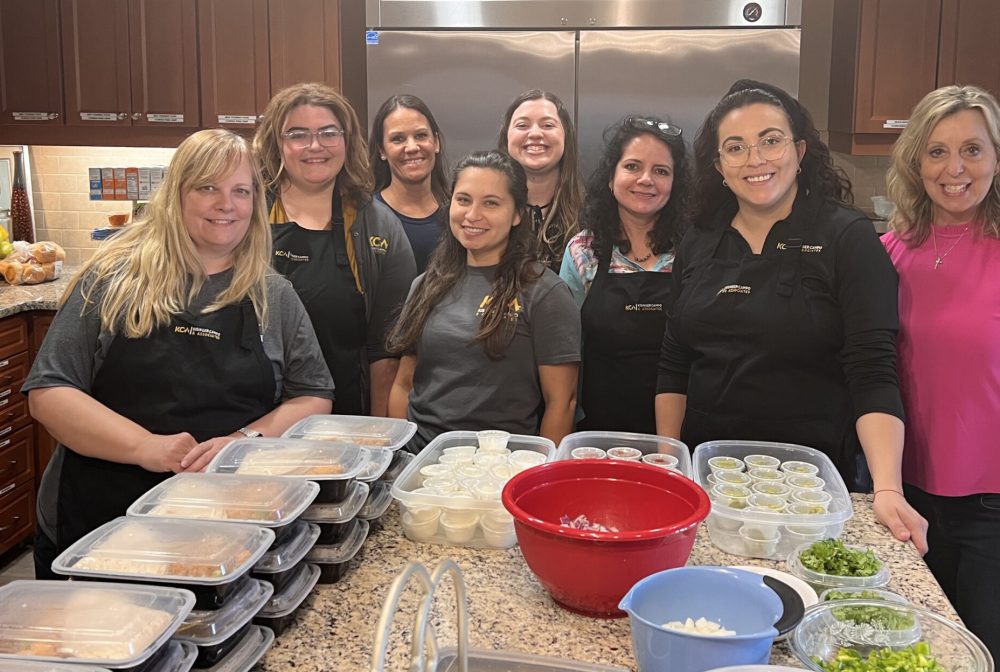 Group of eight women in a kitchen preparing meals, smiling at the camera