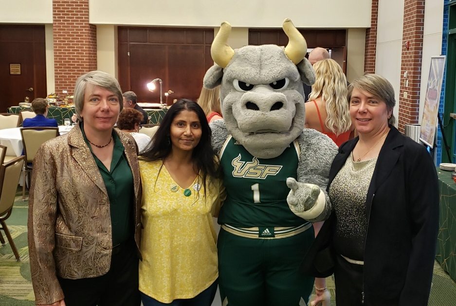 A mascot in USF uniform poses with three women, smiling in a conference room