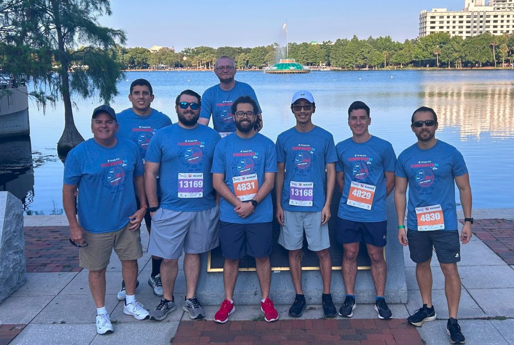 Group of eight men in matching blue shirts posing by a lakeside in Orlando, smiling, wearing race bibs, with cityscape background
