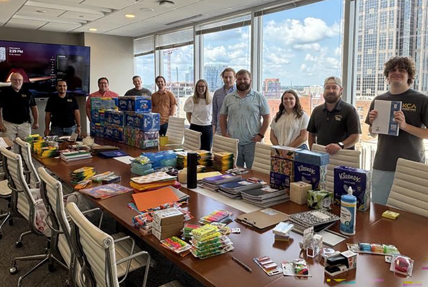 Group of ten people standing behind a conference table filled with school supplies in a bright office with city view