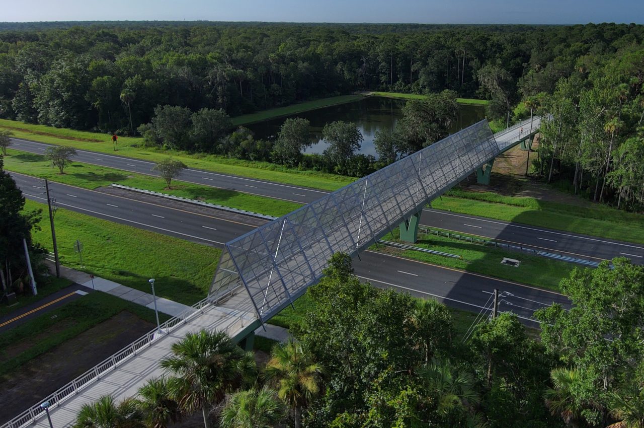 Aerial view of a pedestrian bridge crossing over a road, surrounded by lush greenery and a pond under a blue sky