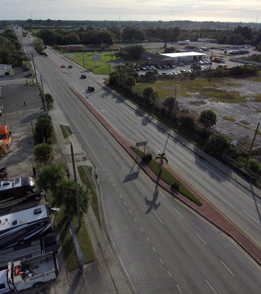 Aerial view of a wide road with scattered vehicles, adjacent commercial buildings, and grassy open land