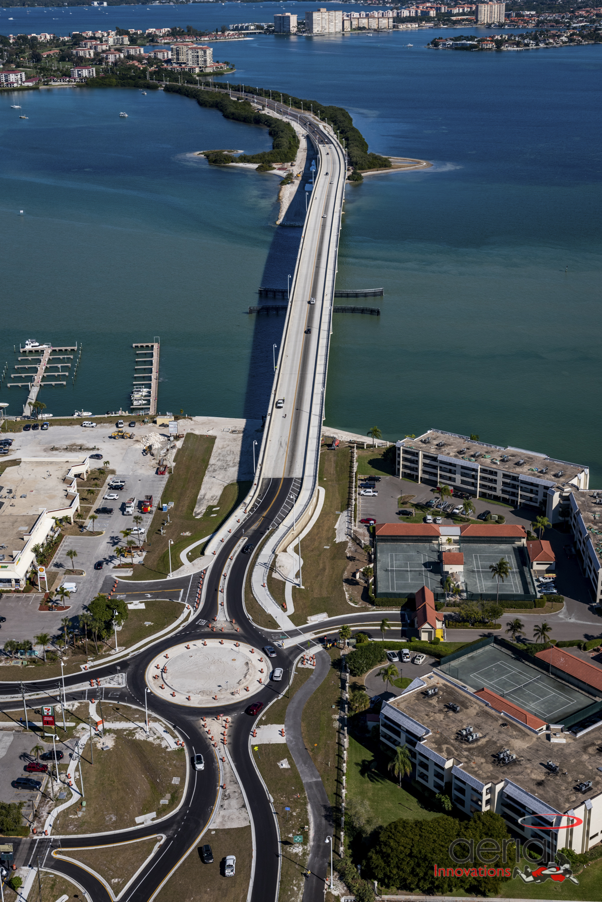 Aerial view of Pinellas Bayway bridge extending over turquoise water, nearby marina and buildings, roundabout in foreground