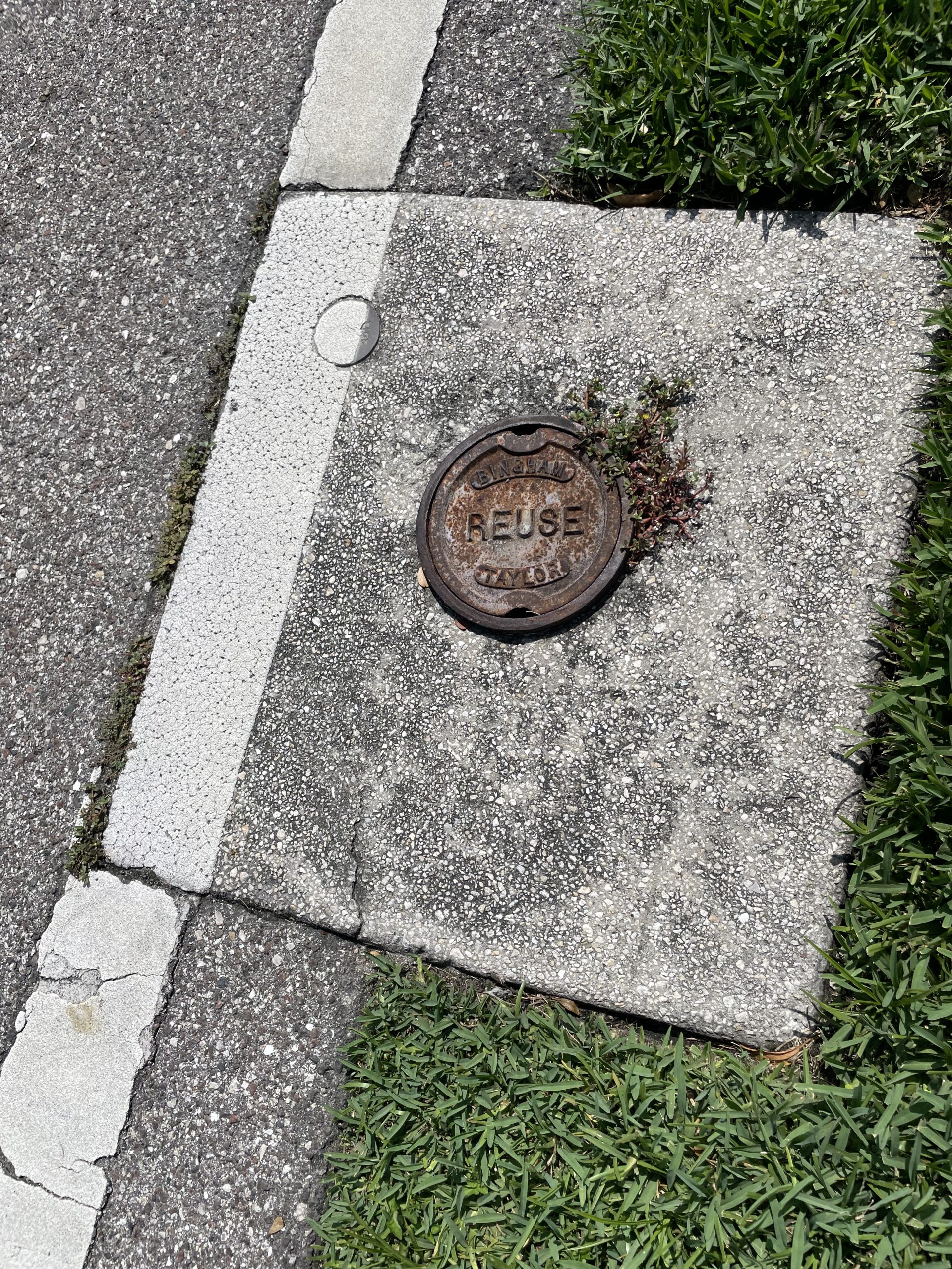 A round metal cover on the ground labeled "REUSE WATER" beside grass and pavement
