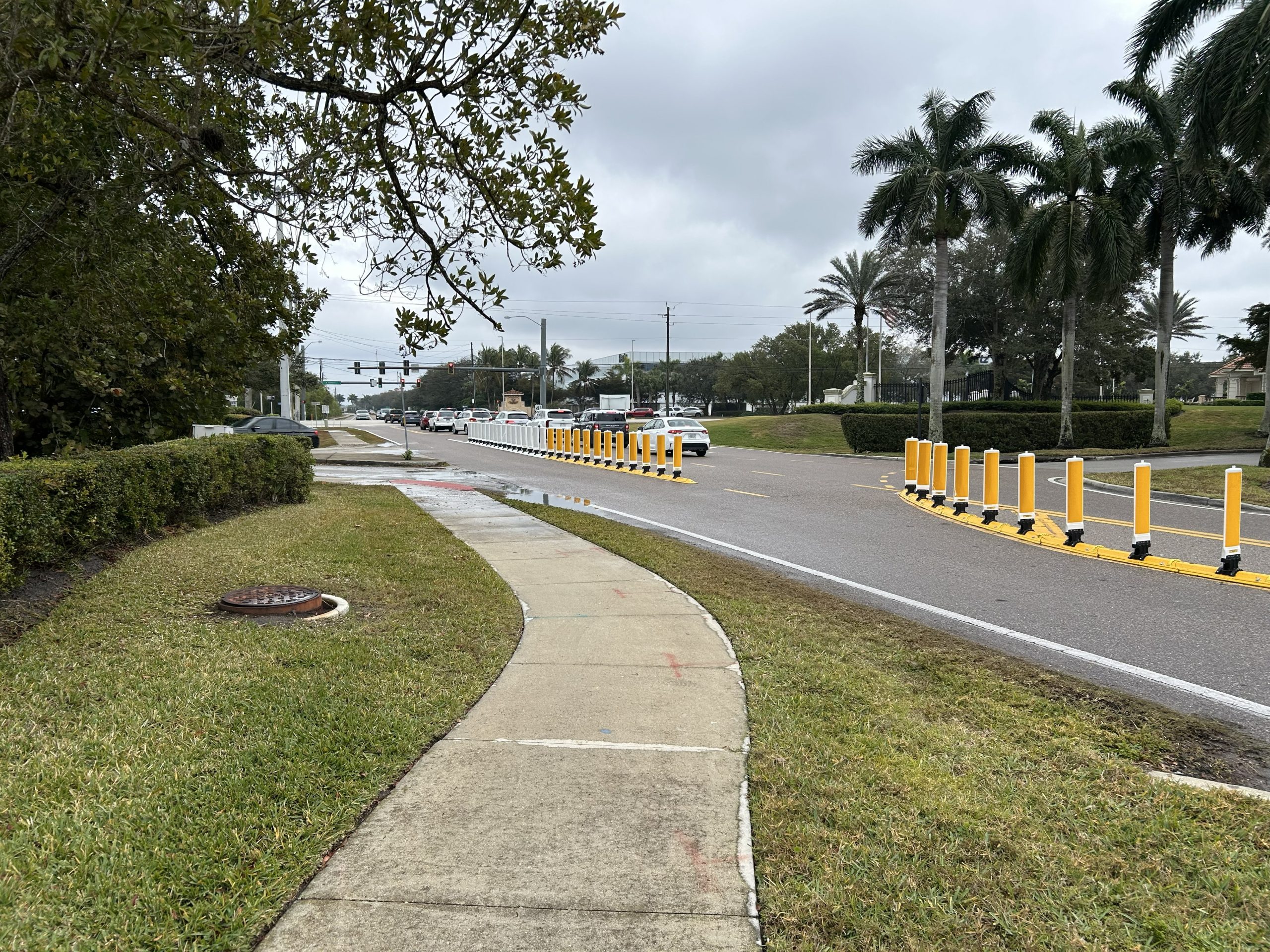 Sidewalk alongside road with yellow traffic delineators, palm trees, and cloudy sky